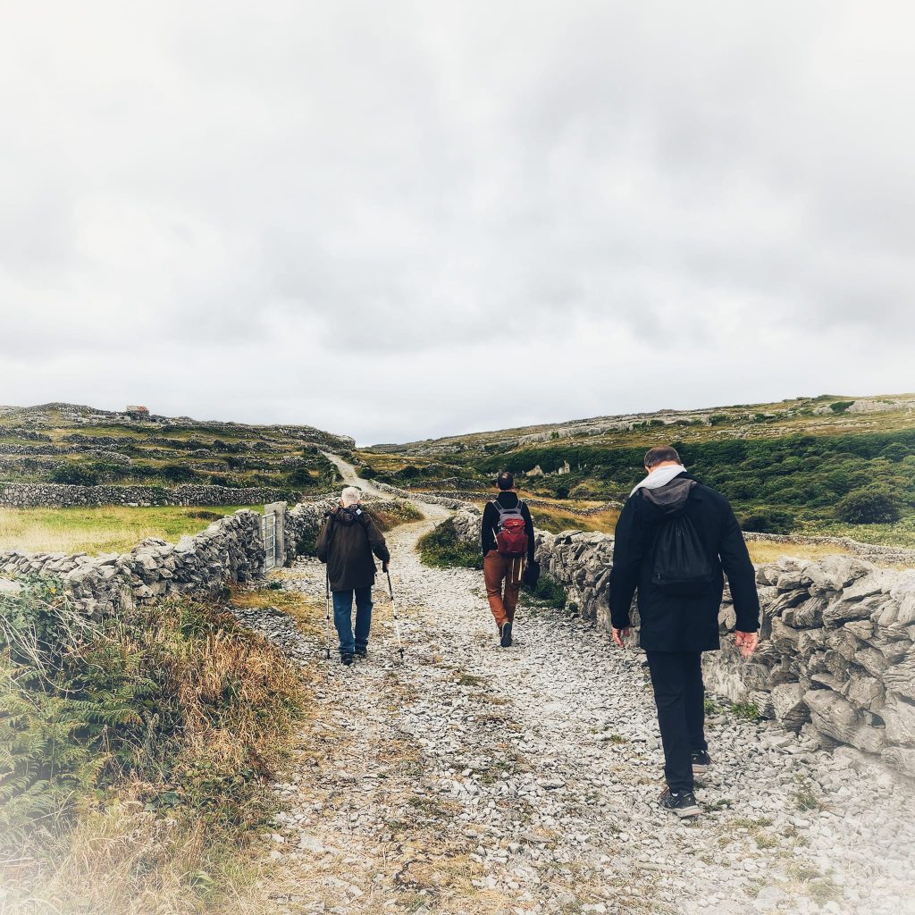 Three of the pilgrims, men, walk ahead of the photographer, They are on a stone path with rock walls on either side. nothing but green grass and stone walls ribboning the sight beyond. Pilgrimage to Ireland.