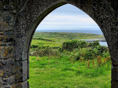 Looking through arched window of ancient church onto green fields, ocean, and sky on pilgrimage to Ireland