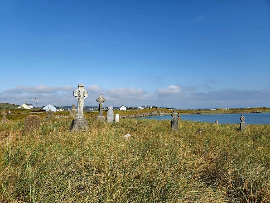 Graveyard in foreground. Headstones capped with tall Irish Celtic crosses. The grass has grown tall and gone to hay. Village, sea, and bright blue sky beyond. Pilgrimage to Ireland.