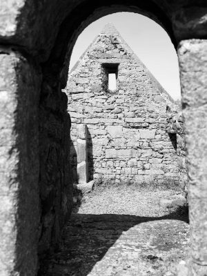 From the outside of an ancient church, looking through an arched window to a wall on the other side. With no roof, the room is lit up by the sunlight. Pilgrimage to Ireland.