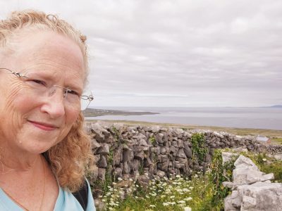 The author, Sarah, smiling at the camera in front of a stone fence with the sea shore, ocean, and sky in the background.