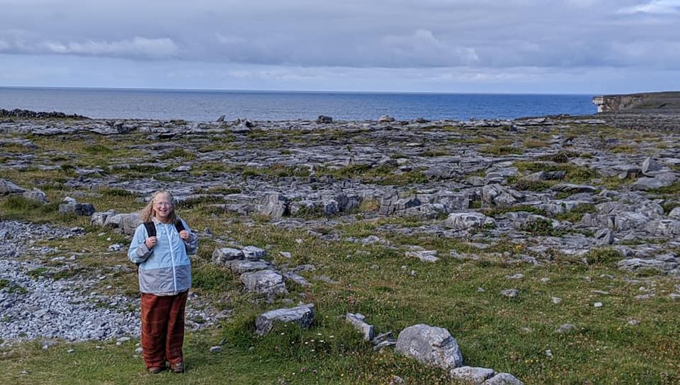 The author, Sarah, standing in a field of grass and large limestone rocks with the ocean and sky in the distance. Pilgrimage to Ireland.