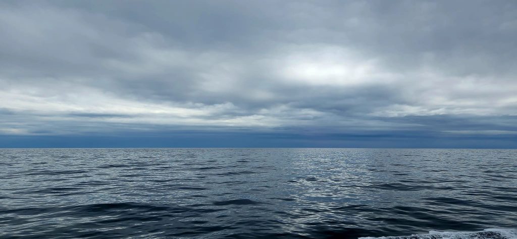 Wide, expansive photo of the Atlantic ocean with a sky full of clouds. The ocean water is dark blue-grey. The clouds are grey in the foreground, some light shines through in the middle, and then the clouds turn a threatening blue at the horizon. 