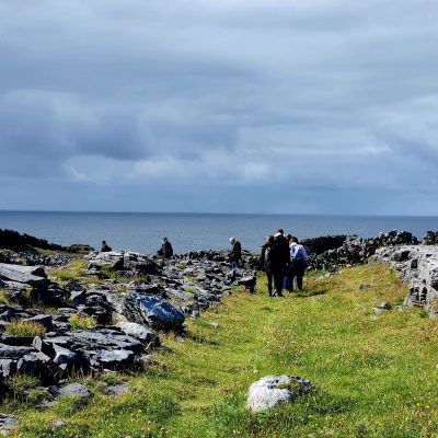 Pilgrims on grassy path with large limestone bolders to either side of the path. Ocean and sky beyond. Pilgrimage to Ireland.