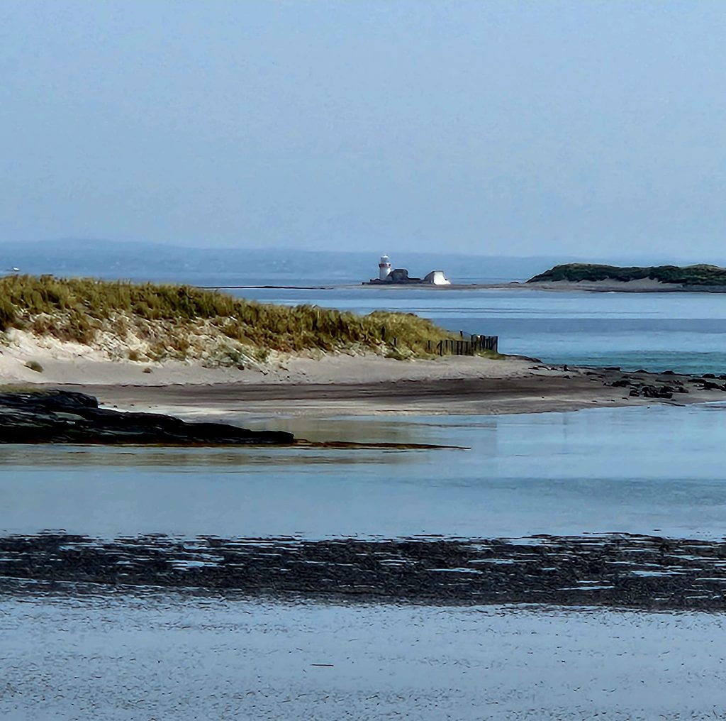 Seashore in foreground with light house in distance and beautiful ocean water all around. 