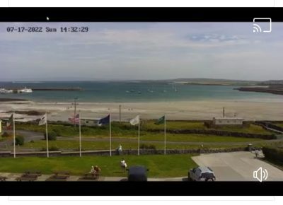 A photo of the video Joseph saw of Sarah standing in front of her hotel. She stands under the six flags with the seashore, ocean, and sky in the background. She is tiny because the camera lens is meant to take in as much of the scenery in front of the hotel as possible. 