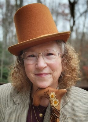 The author, Sarah Elizabeth, in portrait sized photo wearing a brown top hat, a brown checked man's oversized coat, in front of a forest and leaning her chin on the handle of her cane.